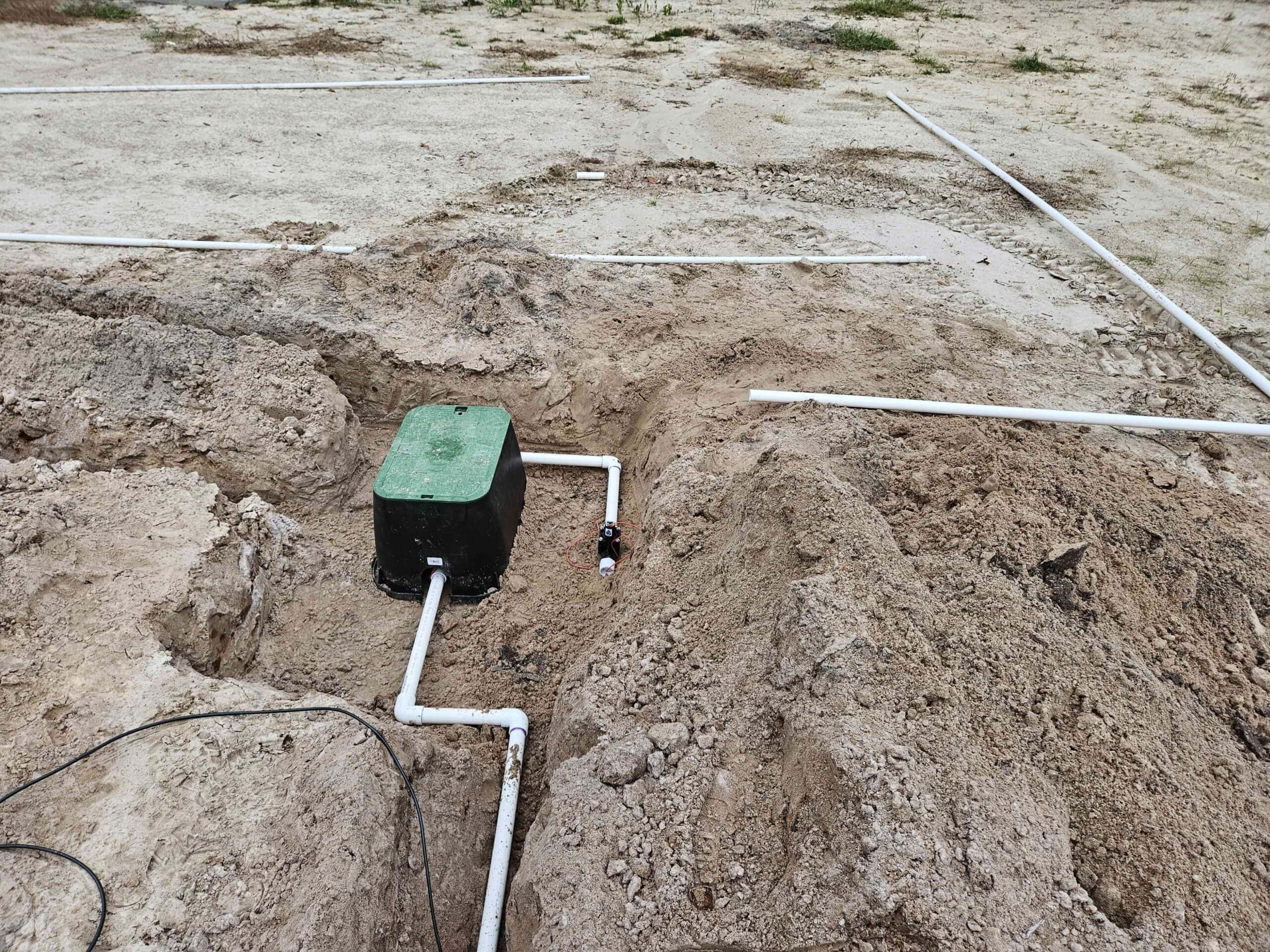 Partially installed underground irrigation system with a green valve box, PVC pipes, and trenches in sandy soil for water distribution.