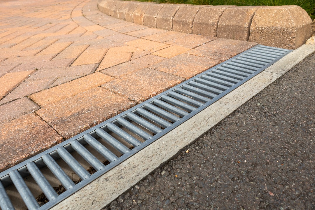 Metal drainage grate along the edge of a paved driveway and asphalt road, designed for water runoff management.