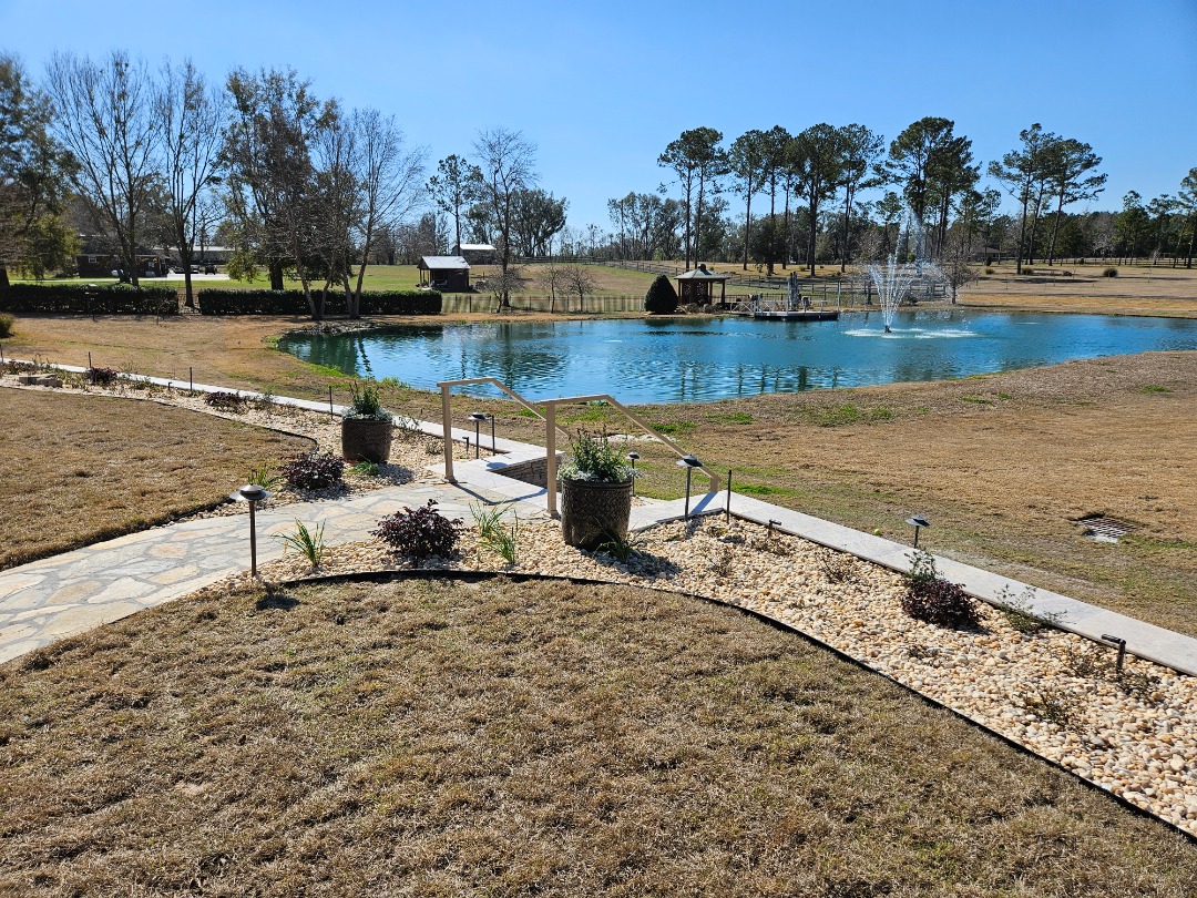 Stone path curves toward a pond with a fountain, bordered by decorative gravel and landscape lighting, set in a rural open field.