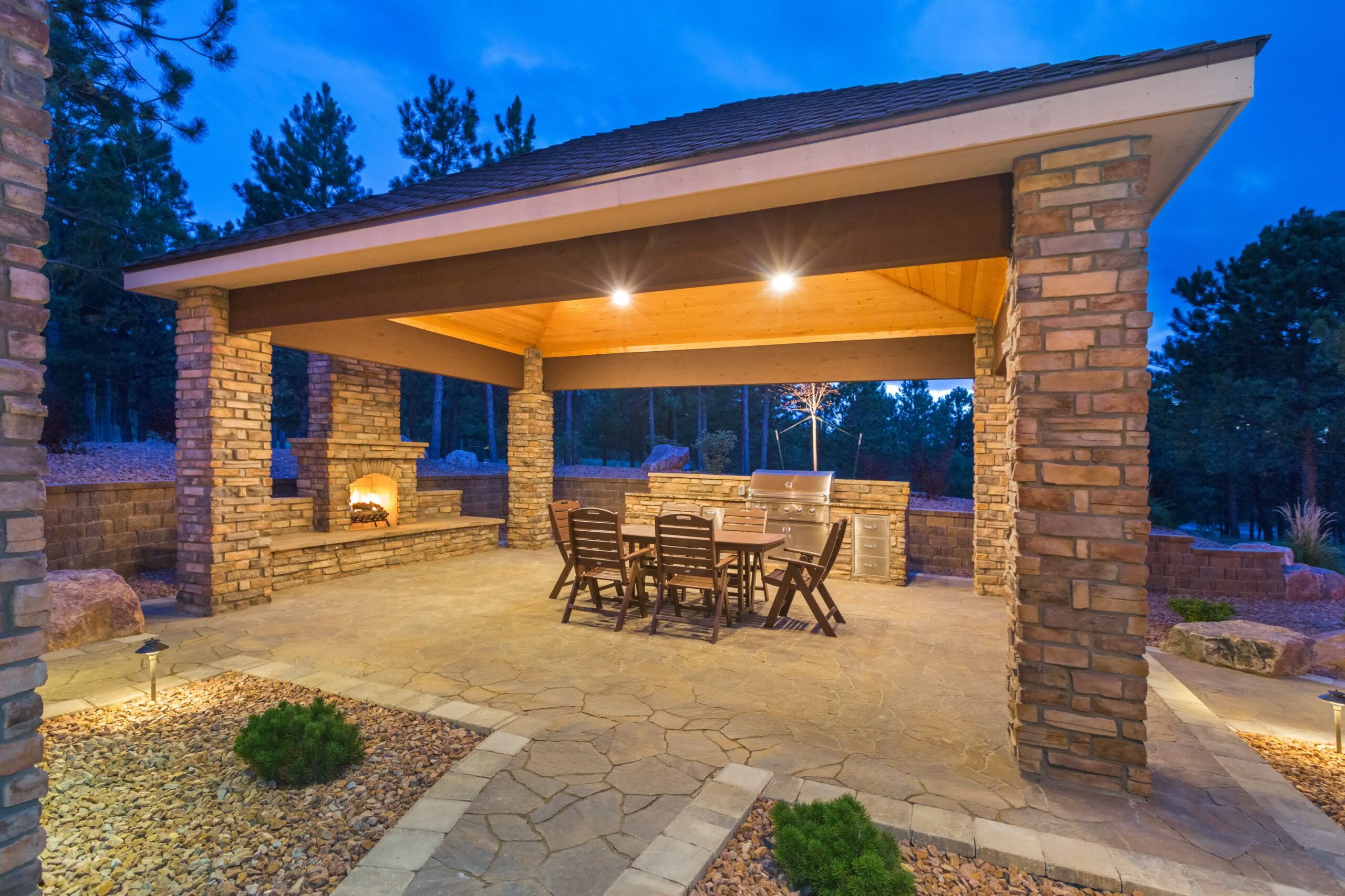 Covered outdoor kitchen with stone fireplace, grill, and dining table sits on paver patio lit warmly during early evening hours.