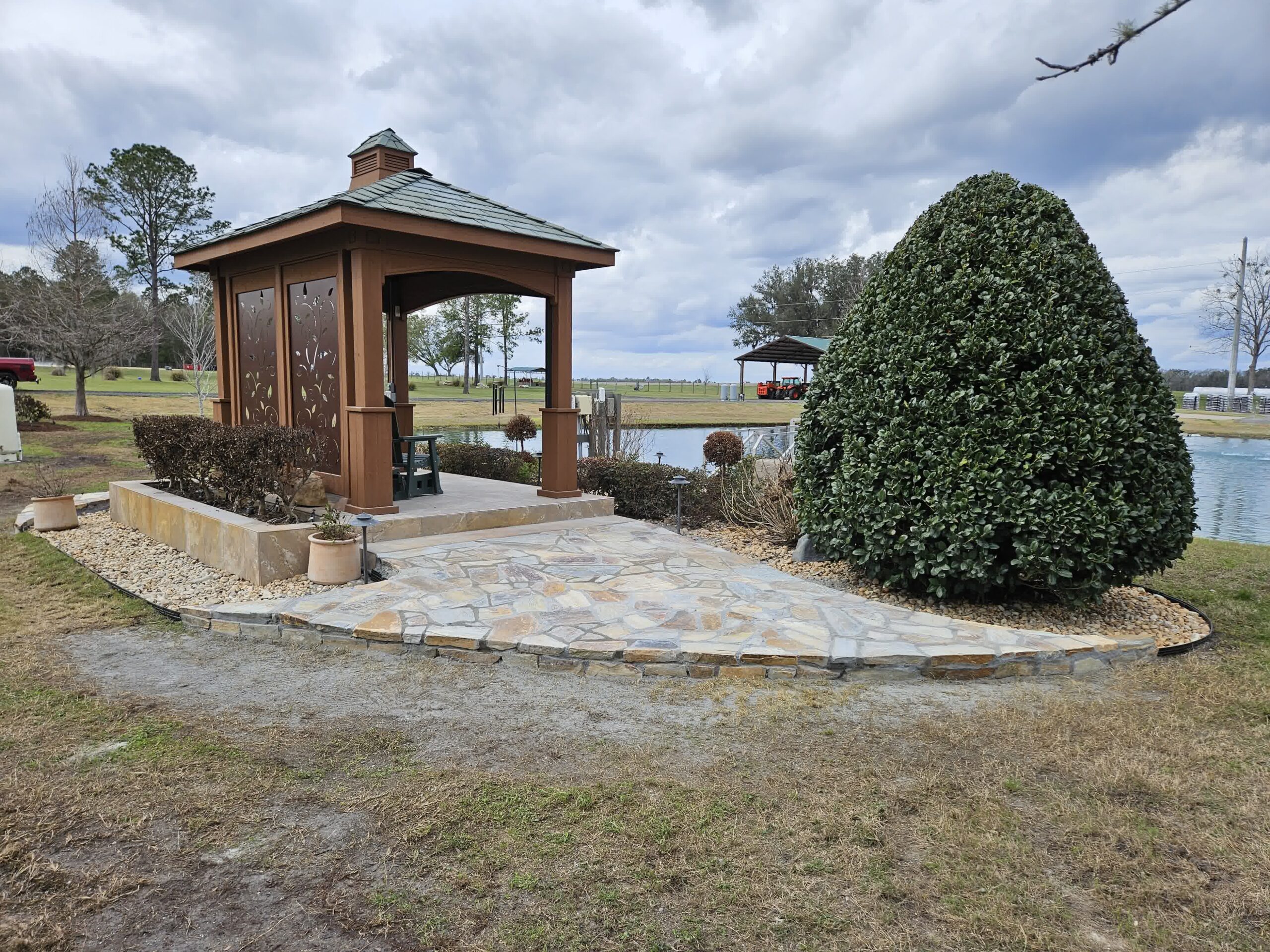 Gazebo with decorative panels sits on stone patio near pond in landscaped yard, surrounded by trimmed shrubs and potted plants.