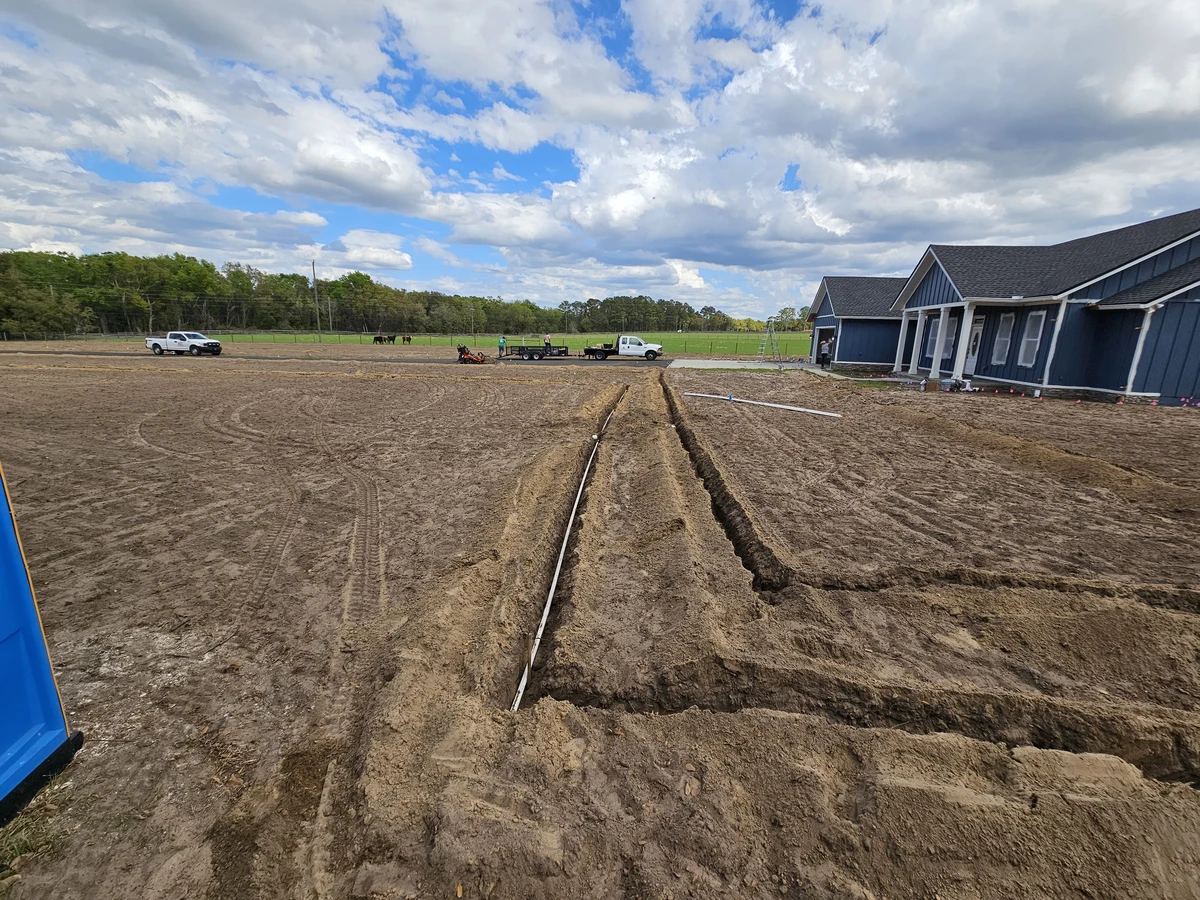 customacreslawn-Irrigation-Ellisvill Irrigation trenches cut through a wide brown dirt yard toward a blue house under a bright cloudy sky.