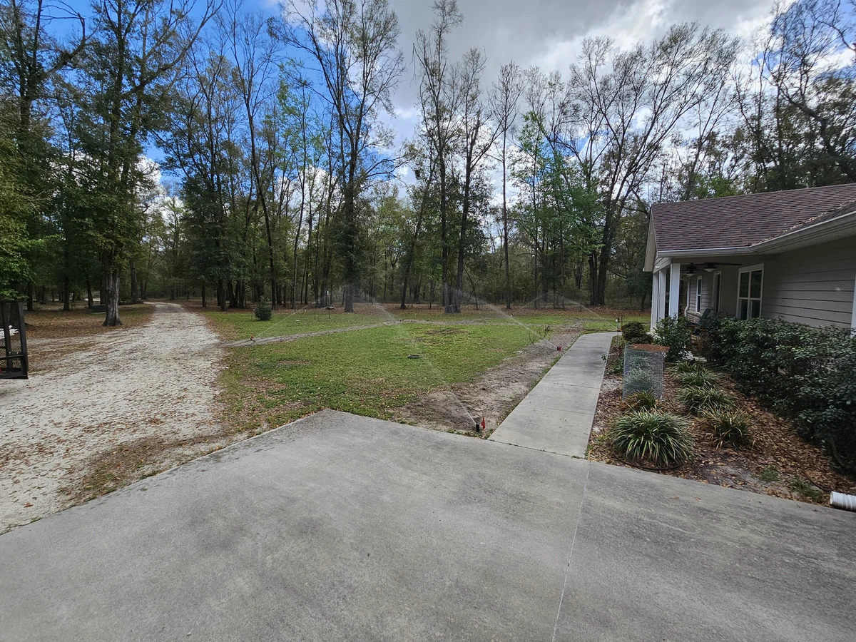 customacreslawn-Irrigation-Lake-City Irrigation lines trace a green yard beside a pale house, with a concrete walkway and tall trees in the background.<br />